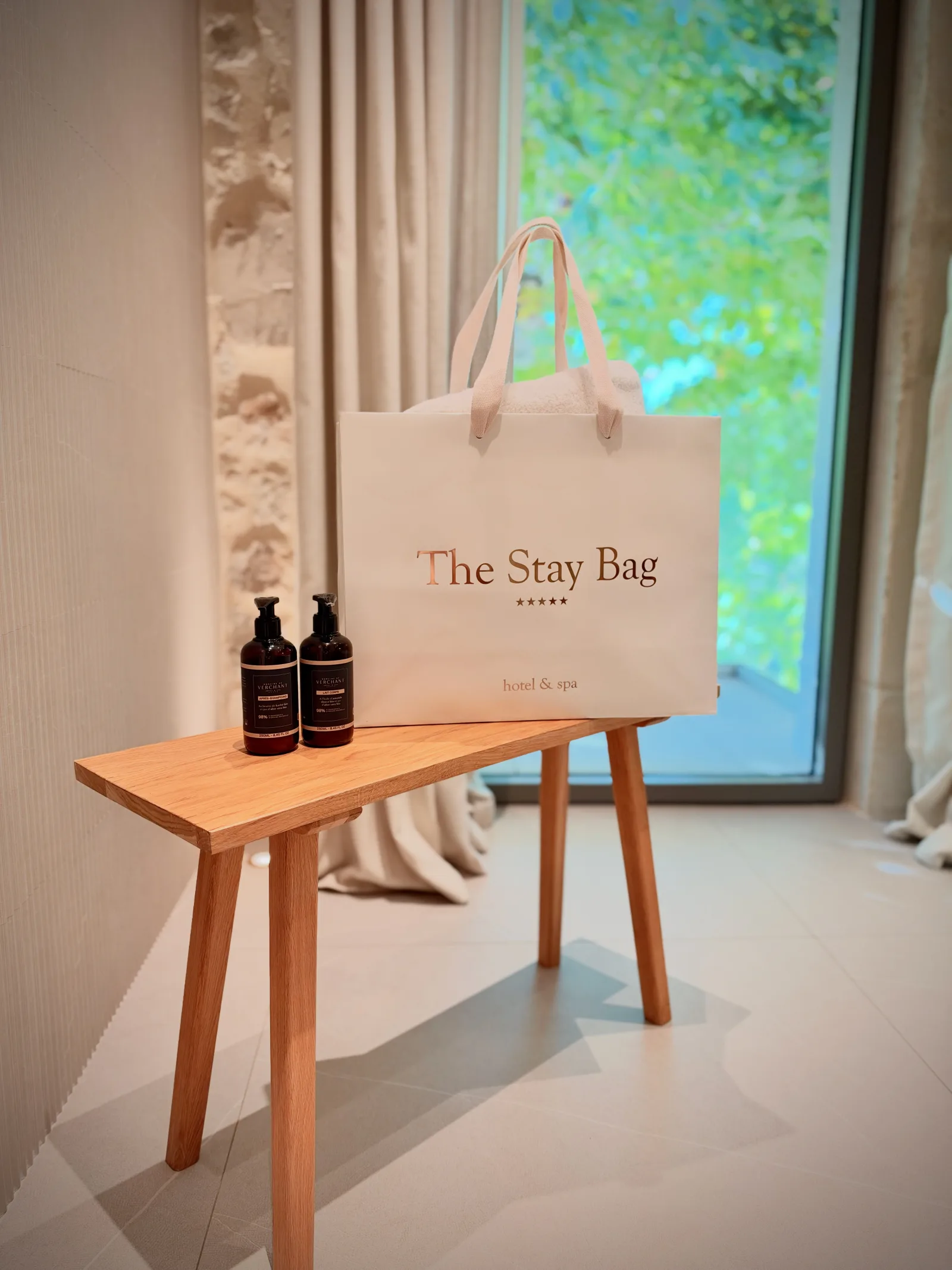 The Stay Bag posé sur un banc en bois, à côté de deux flacons de soin, devant une fenêtre ouverte sur le jardin