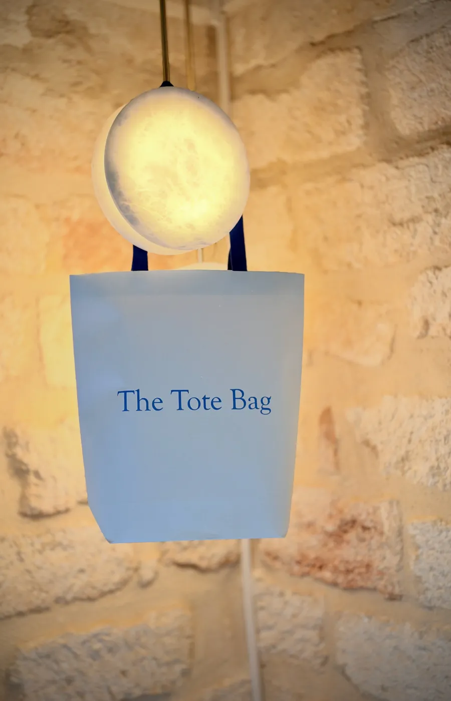 The Tote Bag suspendu par ses anses bleues contre un mur en pierre